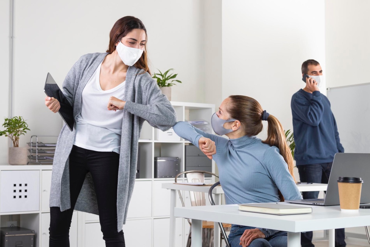 3 people in a bright office wearing face masks due to Covid-19. The two women at the front are bumping elbows.