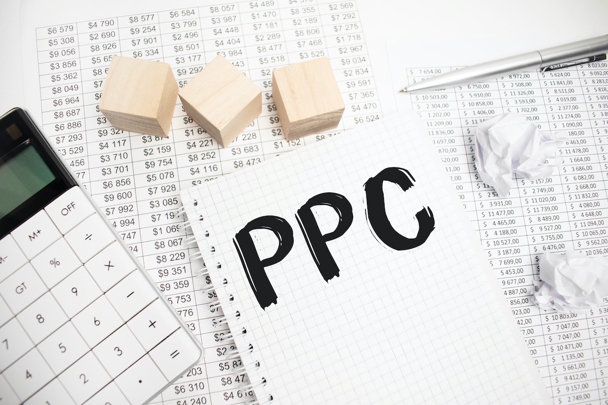 A Spreadsheet, Pen, Calculator, Wooden Blocks, Crumpled Paper, And A Large Notepad In The Middle With “Ppc” Written On It In Black Marker