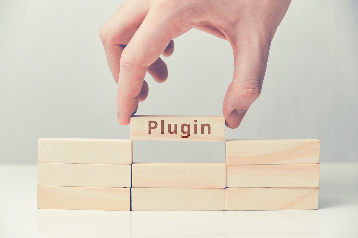A Stack Of 3X3 Wooden Blocks On A White Surface. A Hand Is Reaching Down To Place The Top Middle Block Which Has “Plugin” Written On It Representing Wordpress Plugins