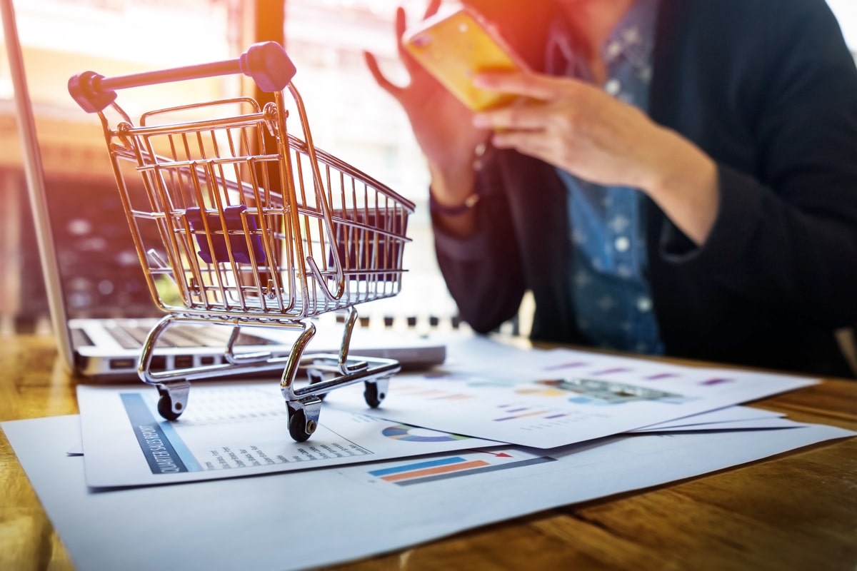 Mini shopping cart placed on papers atop a desk with a laptop, while a woman in the background shops on her phone.