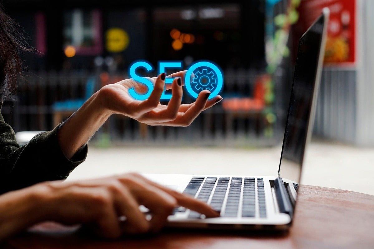 Woman's right hand on a laptop keyboard while another woman's left hand holds illustrated tech-related words like "SEO".