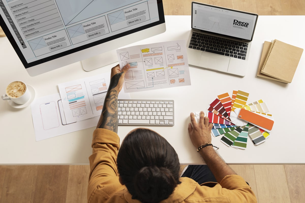 Top view of a web designer working at a desk with webpage sketches, color swatches, and multiple devices displaying a sample website layout